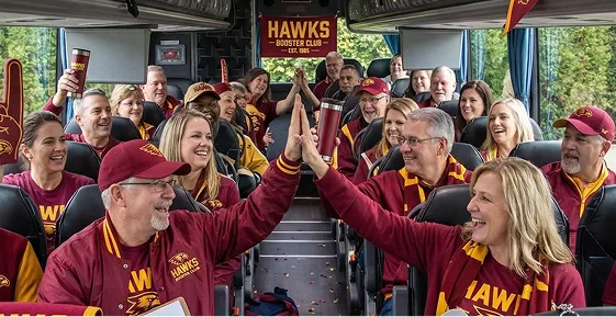 Hawks Booster Club fans in matching maroon jerseys celebrating with high-fives aboard a luxury charter bus