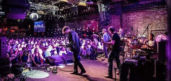 Live band performing on stage before a large crowd in an upscale indoor concert venue with purple lighting