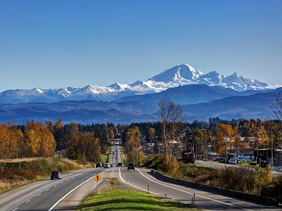 Two-lane Washington State highway in autumn leading toward snow-capped Mount Baker under a clear blue sky