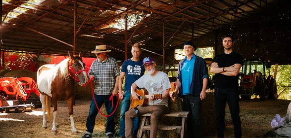 Five men posing in a rustic barn with a paint horse, hay bales, and farm equipment