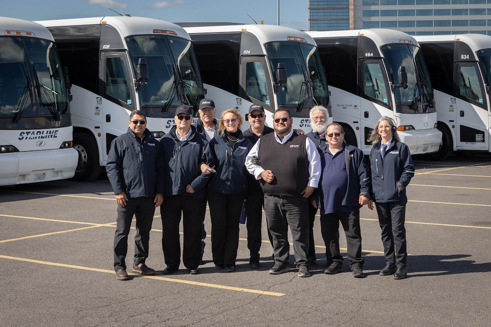 Group of Starline Collection drivers and staff in uniform posed in front of a row of motorcoaches at depot