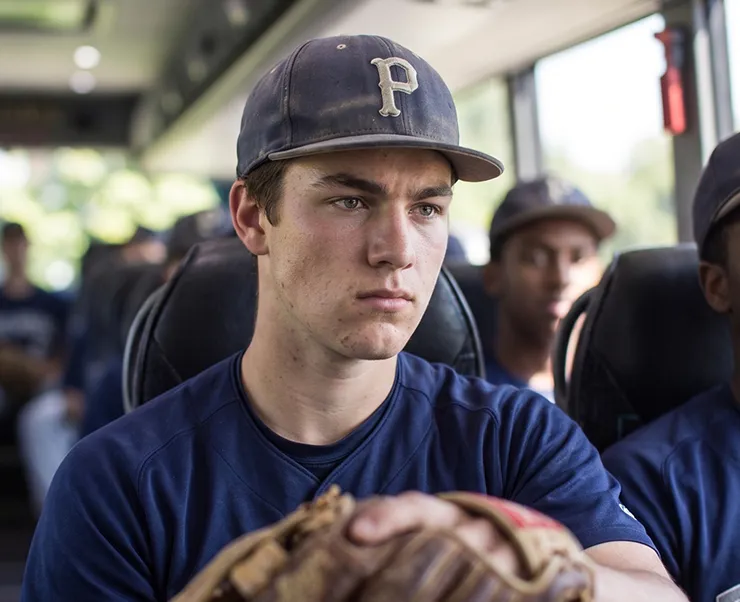 Young baseball player holding a glove seated on a charter bus with teammates in the background
