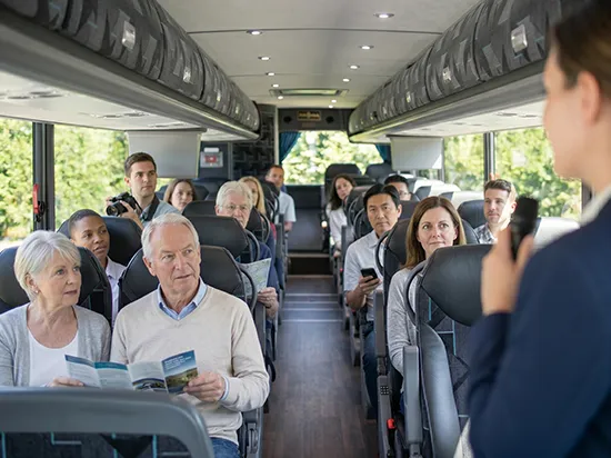 Tour guide addressing seated passengers inside a luxury charter bus with overhead storage and plush seating