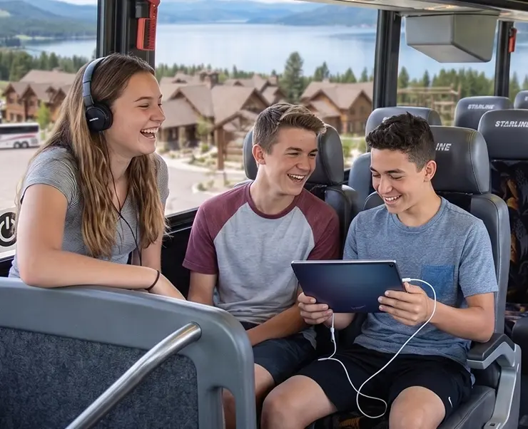 Three teenagers laughing and sharing a tablet aboard a Starline charter bus with scenic lake views