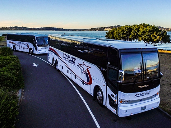Two Starline Collection luxury charter buses parked along a scenic Pacific Northwest waterfront at sunset