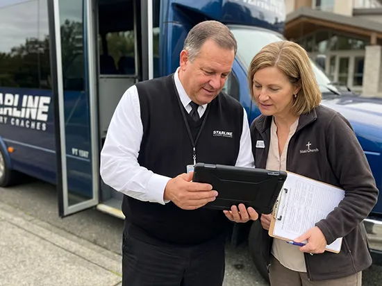 Starline charter bus driver reviewing travel itinerary on tablet with event coordinator near luxury coach