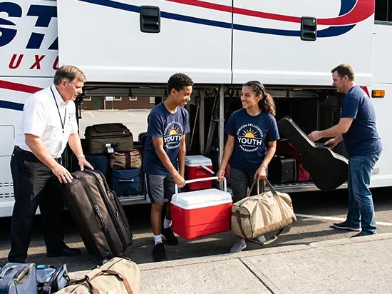 Youth group passengers loading luggage into charter bus undercarriage with driver assistance