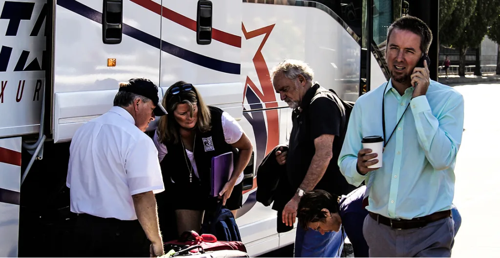 Several people loading luggage outside a charter bus, one man talking on a phone and holding a coffee cup
