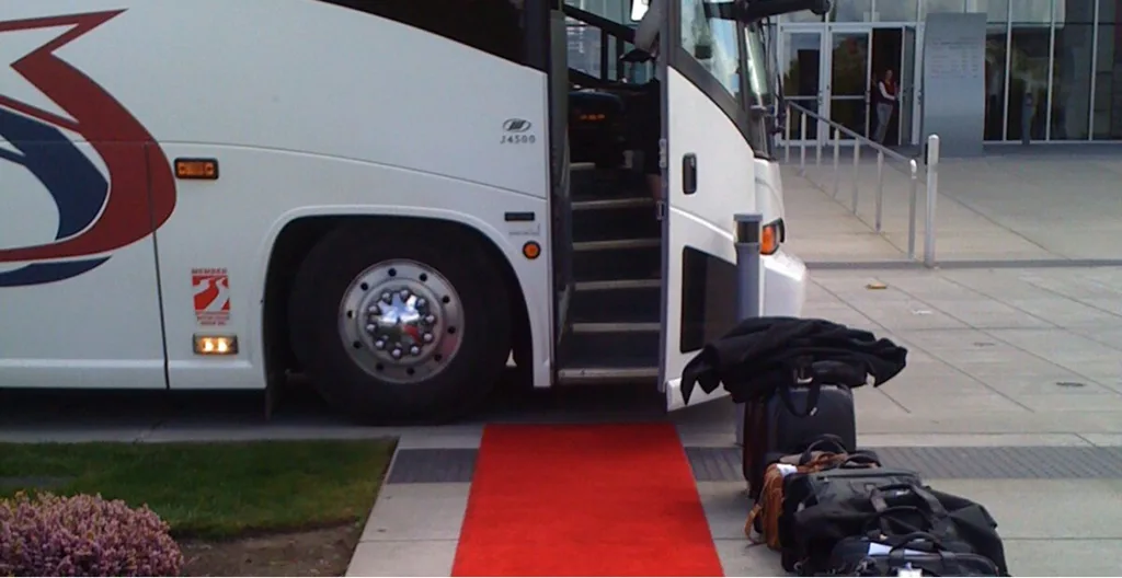 A charter bus with its door open, a red carpet leading to the entrance steps, and luggage on the sidewalk outside a building