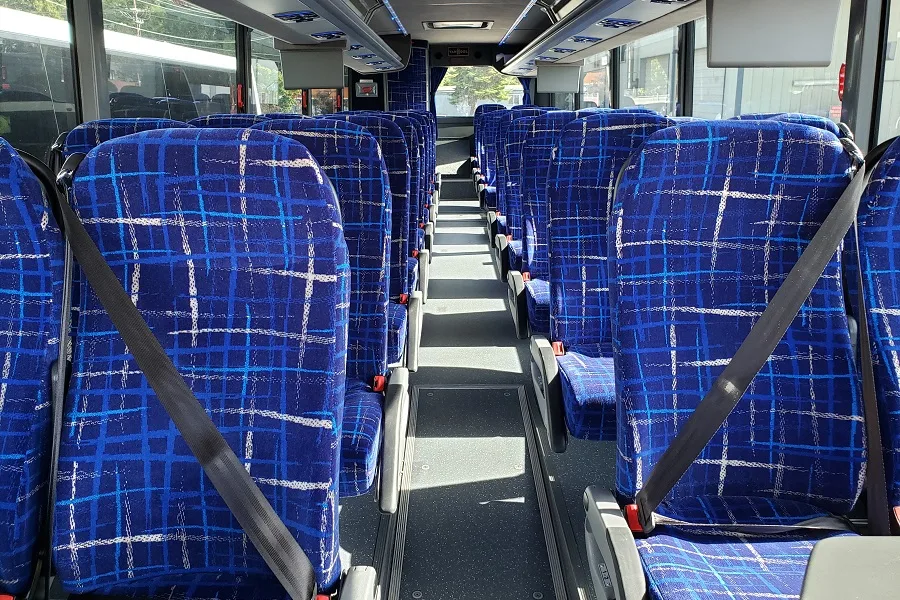 Interior of a charter bus with rows of blue patterned seats equipped with seatbelts and a center aisle