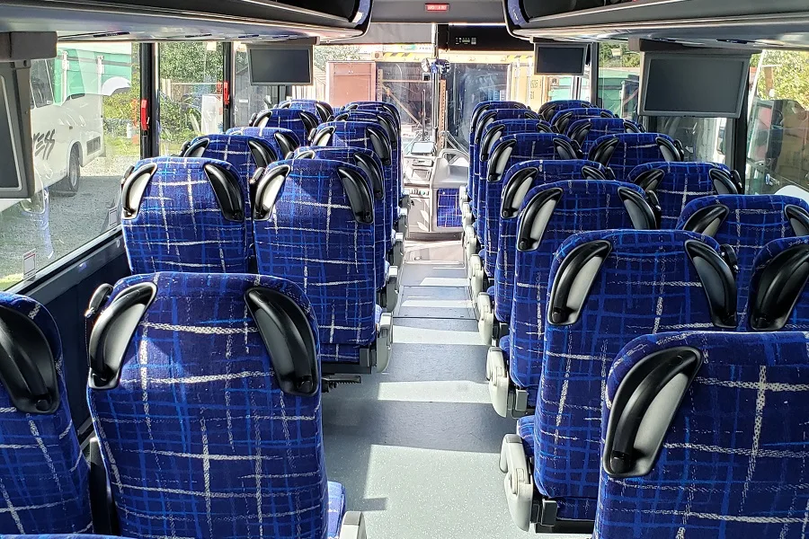 Interior of a charter bus with rows of blue plaid seats, overhead monitors, and a center aisle leading to the driver's area