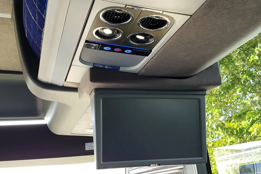 Overhead monitor and climate controls inside a charter bus cabin, showing a ceiling-mounted screen and individual air vents